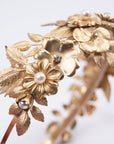 Close-up of THALIA bridal headband on a white surface, showcasing the intricate layers of gold-tone matte brass wildflowers, heritage leaves, and Preciosa pearl centers.