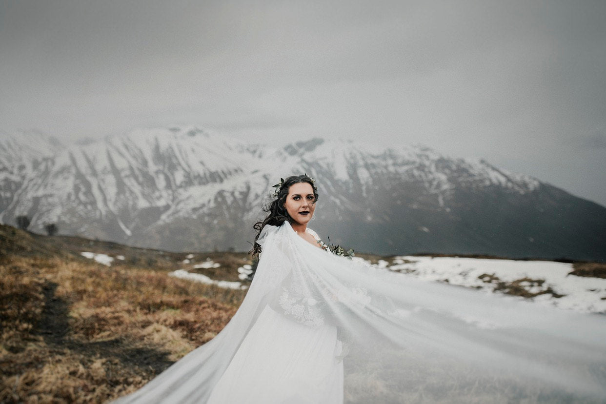 Bride wearing a flowing lace cape veil on a mountaintop elopement, snow-covered peaks in the background.