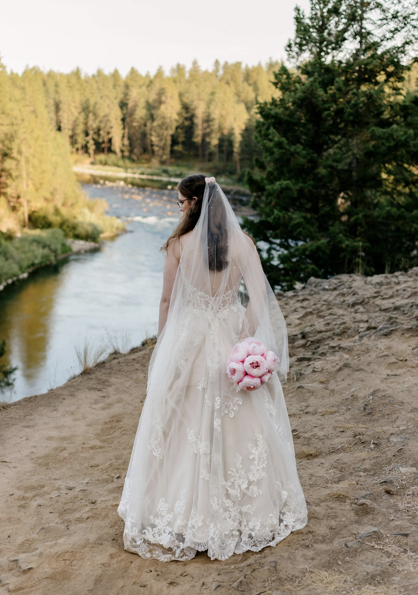 Bride overlooking river after their small forest wedding ceremony, veil softly draping behind.
