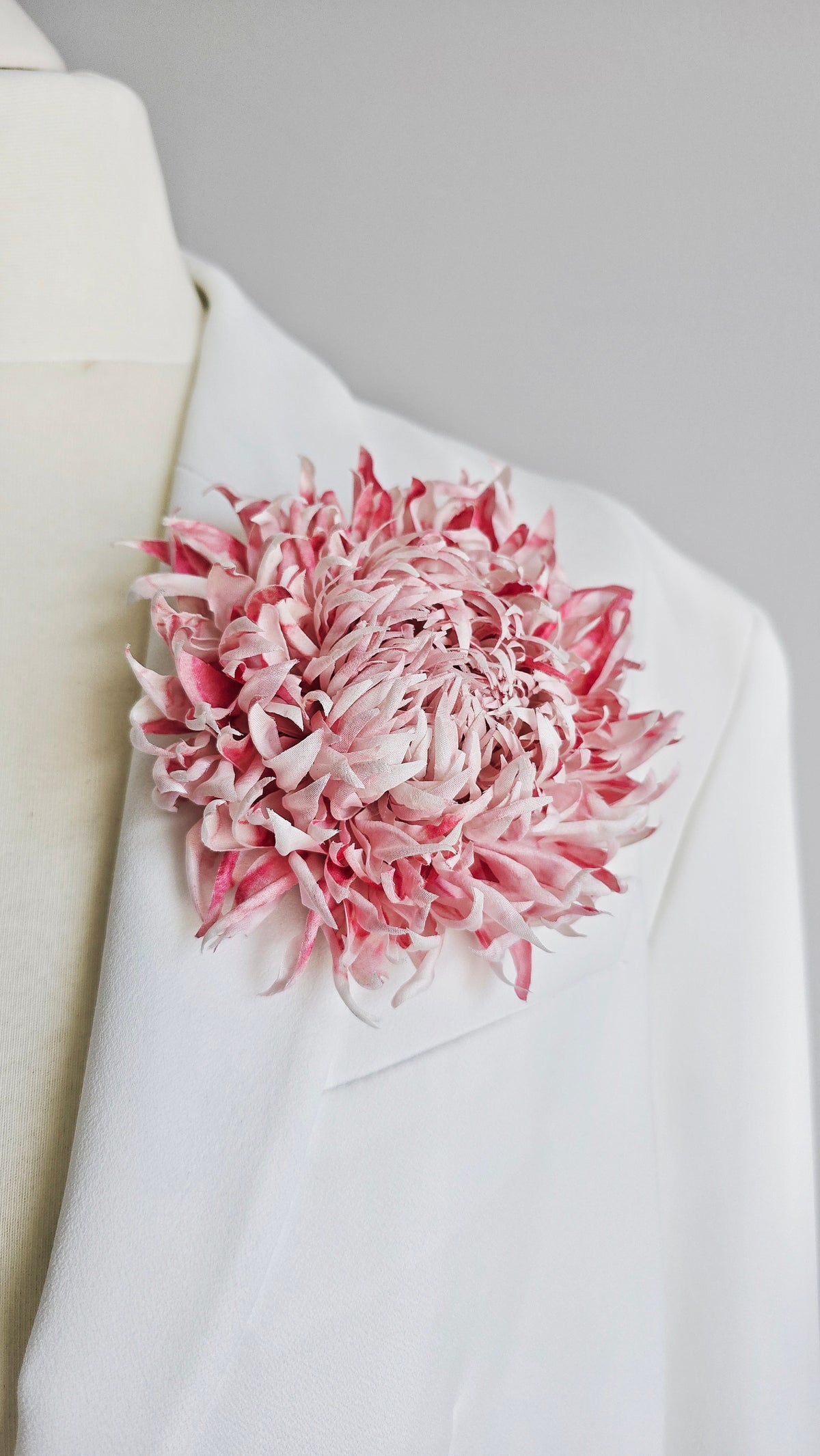 A large, textured pink and soft white silk flower brooch pinned to the lapel of a white suit jacket on a mannequin torso.