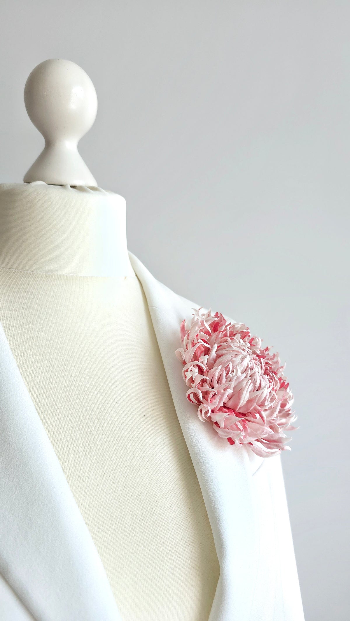 A pink and soft white flower brooch pinned to the lapel of a white blazer displayed on a cream-colored mannequin torso, set against a neutral light gray background.