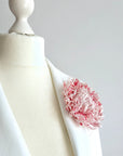 A pink and soft white flower brooch pinned to the lapel of a white blazer displayed on a cream-colored mannequin torso, set against a neutral light gray background.