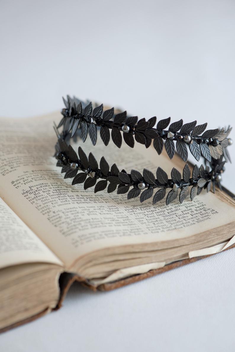 Top-down perspective of the MARIA black laurel leaf crown resting on the aged pages of an open vintage book, highlighting the symmetrical double-row design and matte metallic texture.