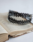 Top-down perspective of the MARIA black laurel leaf crown resting on the aged pages of an open vintage book, highlighting the symmetrical double-row design and matte metallic texture.