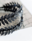 Close-up of MARIA black laurel leaf tiara resting on an antique open book, showing the matte black brass leaves and dark pearls.