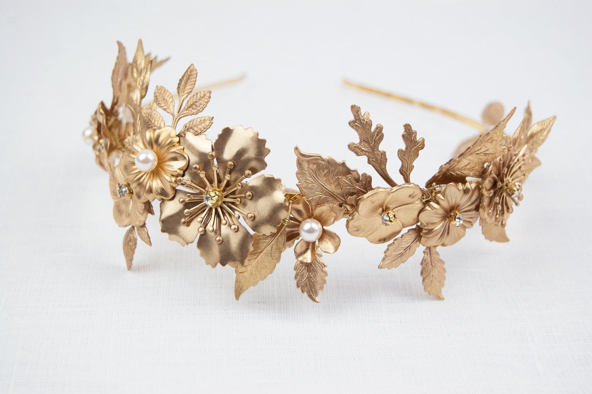 Close-up of THALIA bridal headband on a white linen surface, showcasing the intricate layers of gold-tone matte brass wildflowers, heritage leaves, and Preciosa pearl centers.