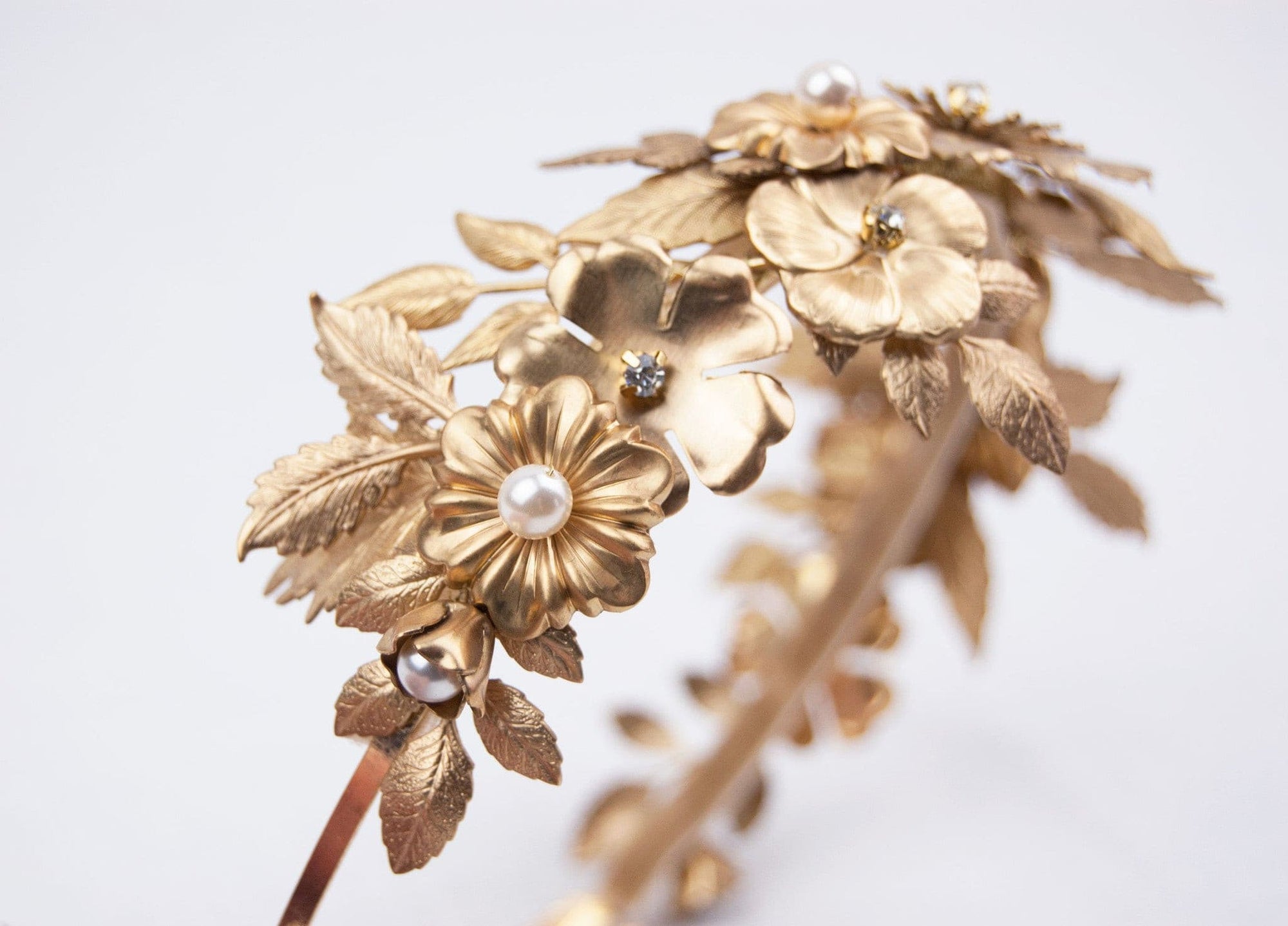 Close-up of THALIA bridal headband on a white surface, showcasing the intricate layers of gold-tone matte brass wildflowers, heritage leaves, and Preciosa pearl centers.