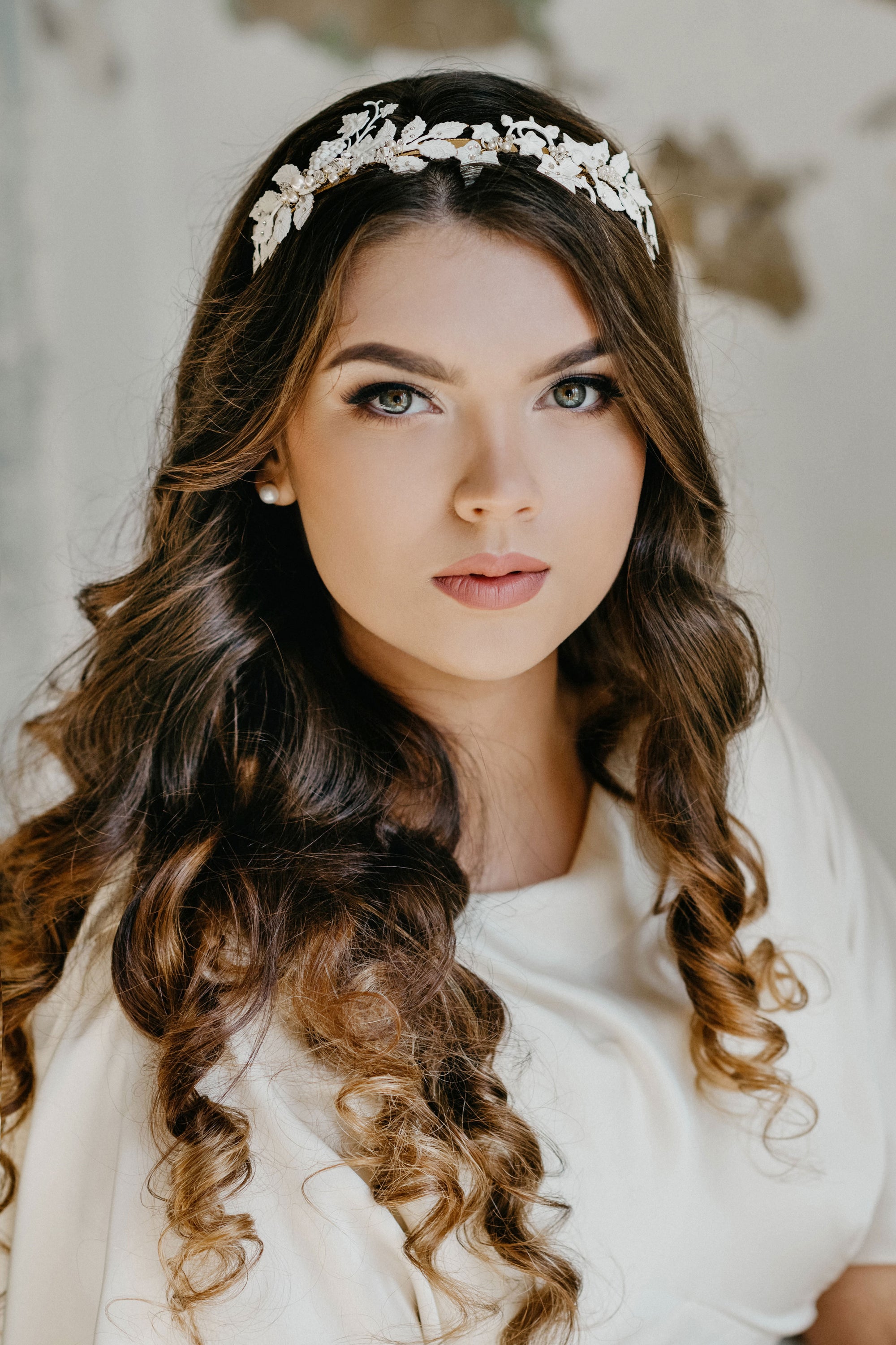 Victoria hand-painted white leaf bridal tiara on a brunette bride with long wavy hair, showcasing the sculptural vine silhouette and matte botanical finish against a textured wall background.