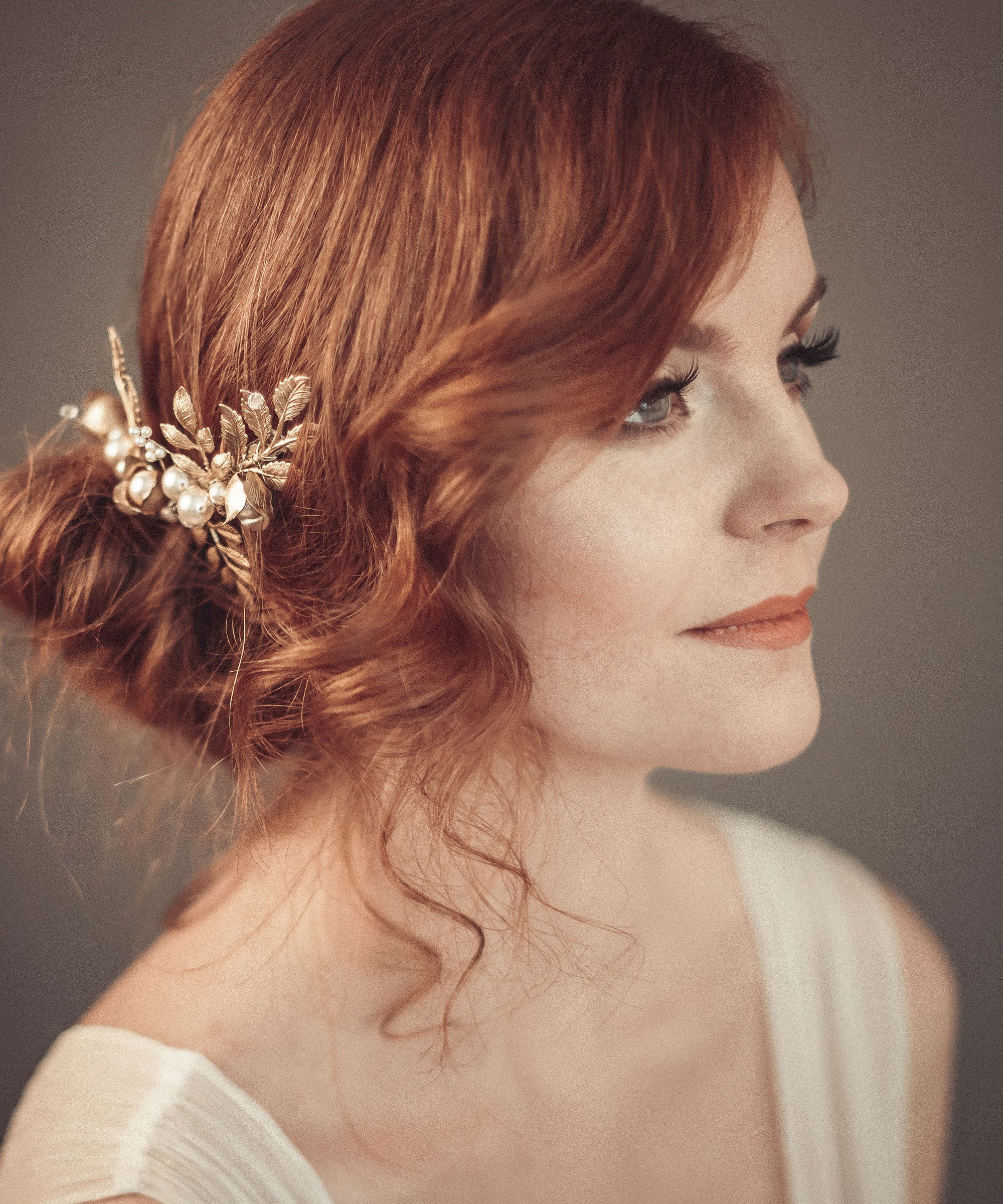 Side profile of a bride with red hair and a low bun wearing the TYCHE gold leaf hair vine comb with pearls for a vintage-inspired bridal look.