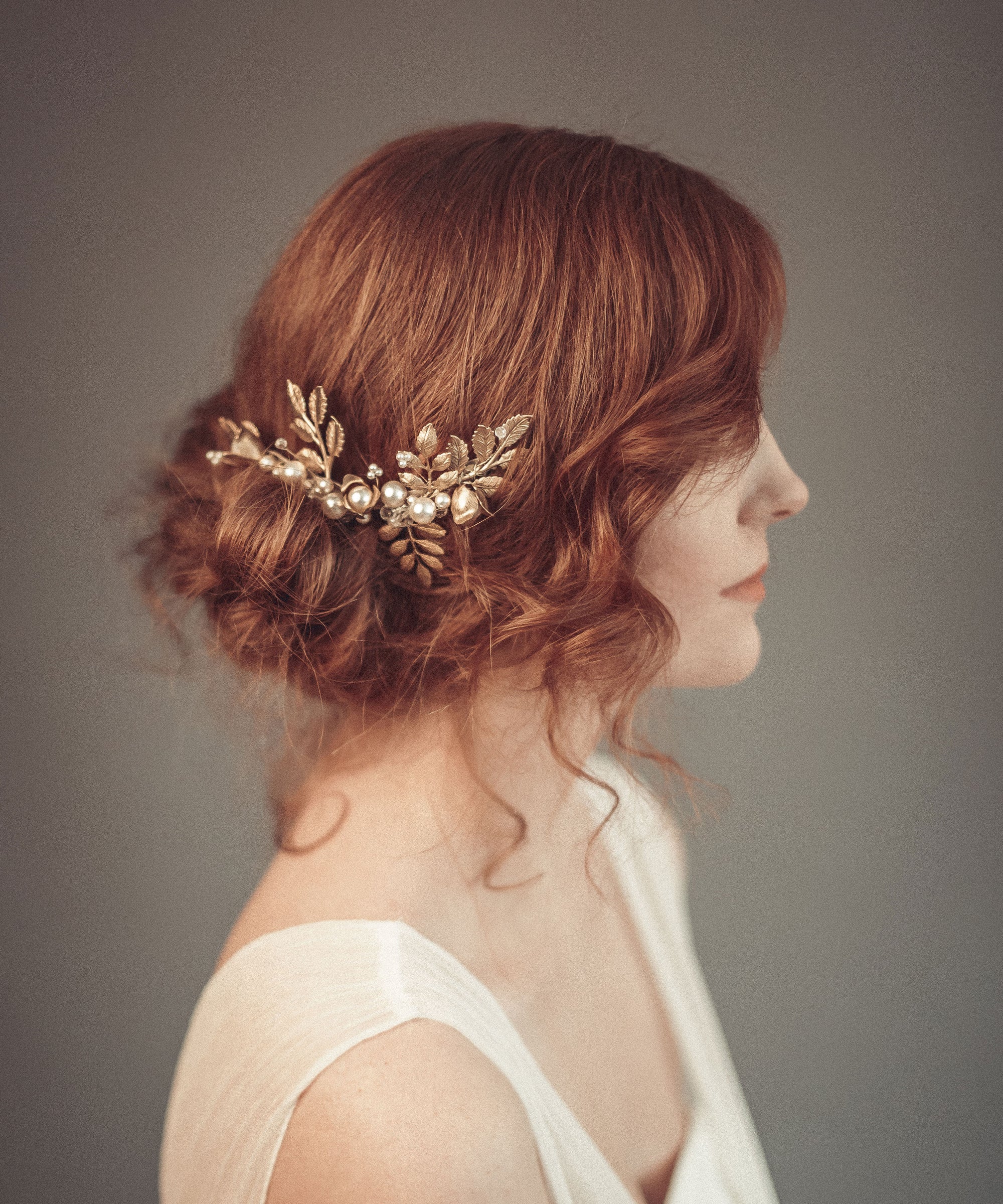 Side profile of a bride with red hair and a low bun wearing the TYCHE gold leaf hair vine comb with pearls for a vintage-inspired bridal look.