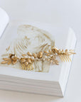 TYCHE bridal hair vine comb with gold brass leaves and pearls resting on an art book, showing the intricate sculptural floral design.